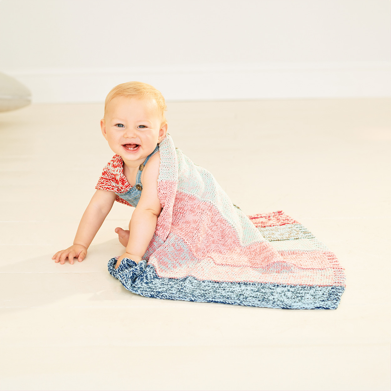 Lachendes Baby sitzt unter einer bunten gestrickten Decke. Fotosetting heller Boden.