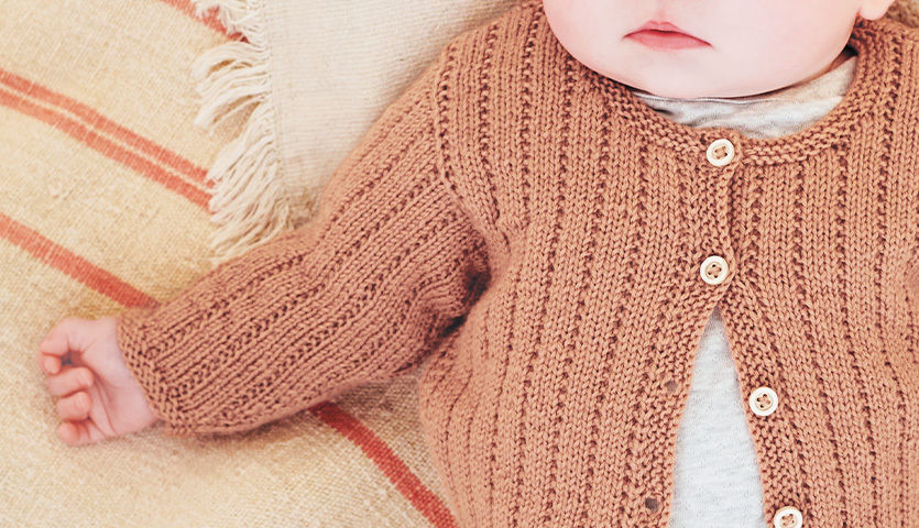 Strickanleitung Jacke aus Baby Merino: Ein Baby trägt eine gestrickte Jacke in sanftem Braunton mit Knöpfen, auf einem gestreiften Kissen liegend.