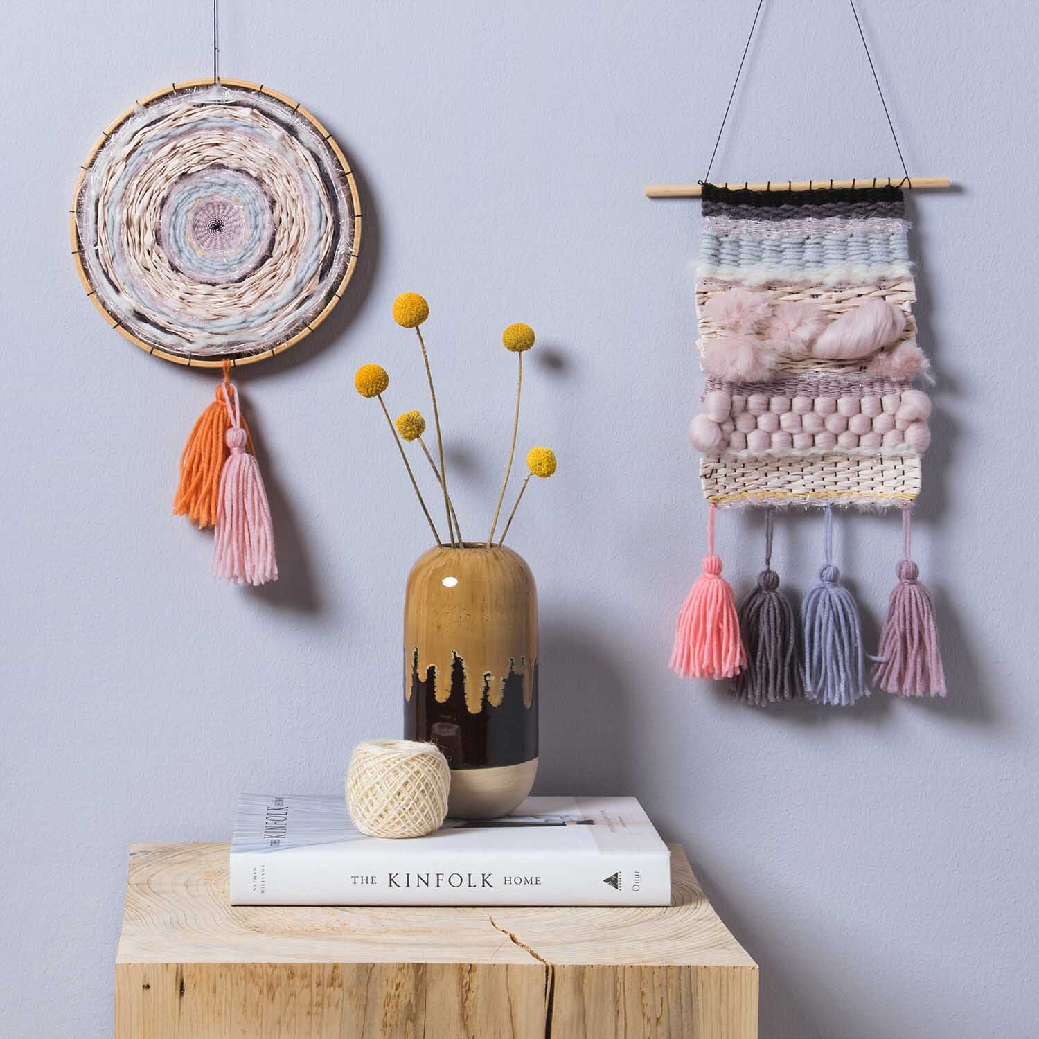 DIY Set Weben: Wanddekoration mit gewebten Mustern und Quasten in Pastelltönen, neben einer Vase mit gelben Blumen.