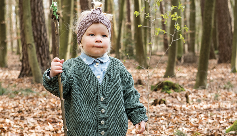 Strickanleitung Kinderjacke und Stirnband aus Essentials Merino dk: Kind im Wald mit grüner Jacke und grauem Stirnband.
