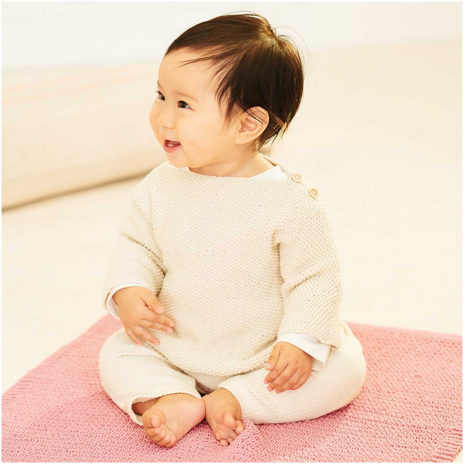 Baby Organic Cotton: Ein lächelndes Baby sitzt auf einer altrosa Decke, gekleidet in einem cremefarbenen Strampler.