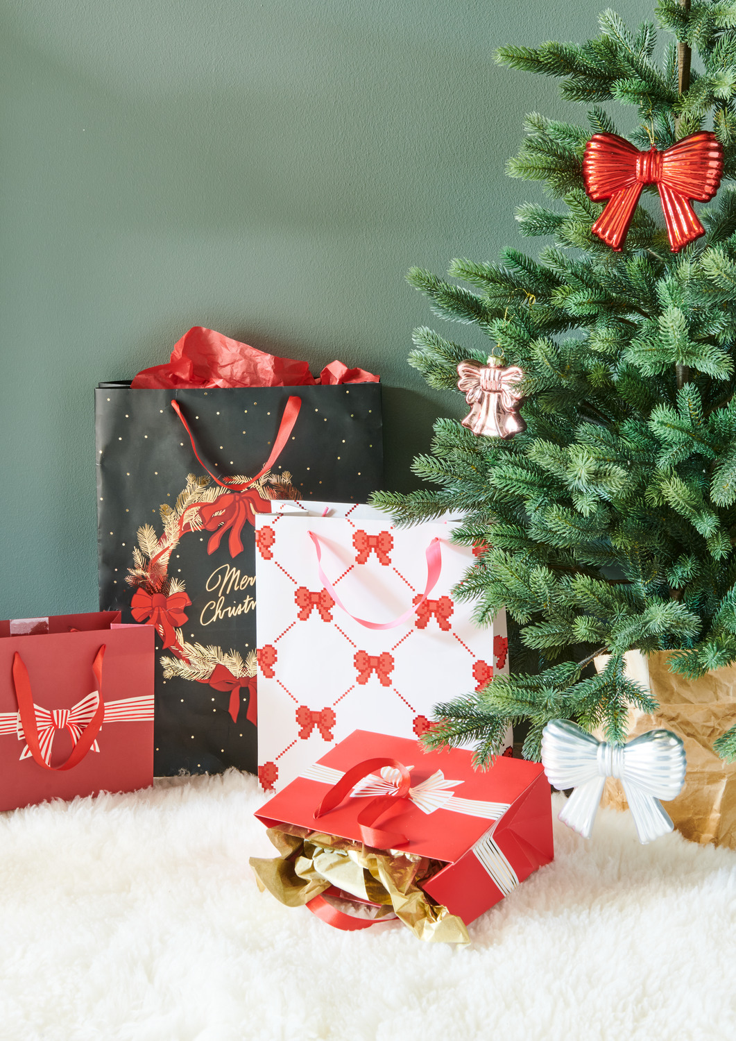 Geschenktüte Pixel Schleifen Creme L neben einem Weihnachtsbaum mit roten und weißen Schleifen, umgeben von weiteren festlichen Geschenktüten.