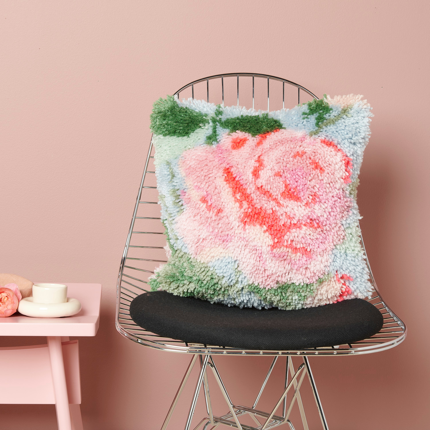 Rico Hooki Hooki Knüpfset Kissen Rose auf einem Stuhl, zeigt ein florales Design in Rosa und Grün. Ideal für stilvolle Wohnräume.