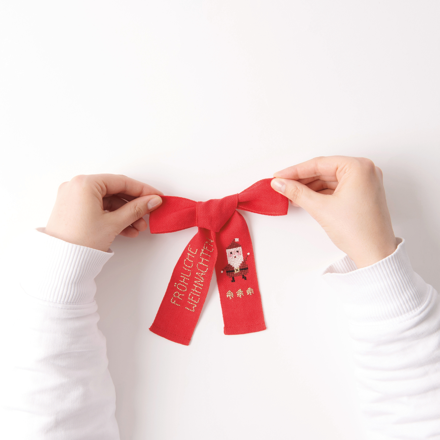 Stickbuch Put a Bow on It Nr. 185: Rote Schleife mit Nikolausmotiv und Schriftzug "Fröhliche Weihnachten", gehalten von Händen.
