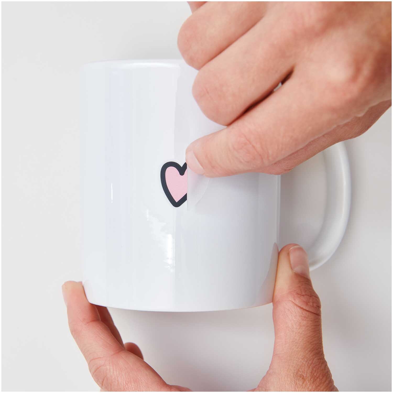 Transfermotiv Just Married 10 Stück: Hände kleben ein Herzmotiv auf eine weiße Tasse. Ideal für Hochzeiten. Marke: Rico Design.