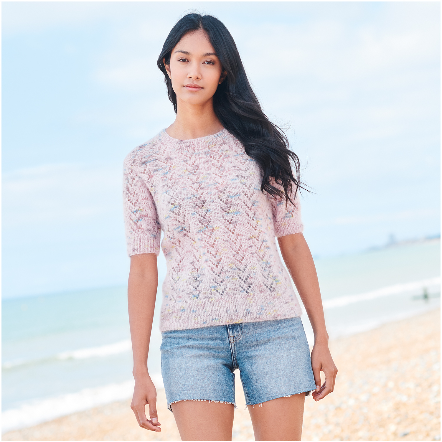Strickset Shirt Modell 12 aus Lovewool Nr. 18: Frau am Strand in hellem Strickshirt und Jeansshorts.