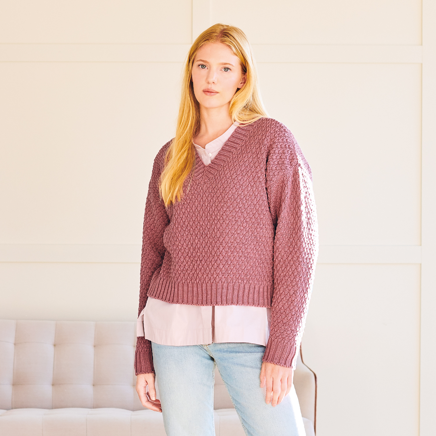 Strickanleitung Pullover aus Essentials Merino dk: Frau trägt gestrickten Pullover in Rosa, kombiniert mit heller Bluse und Jeans, vor heller Wand.