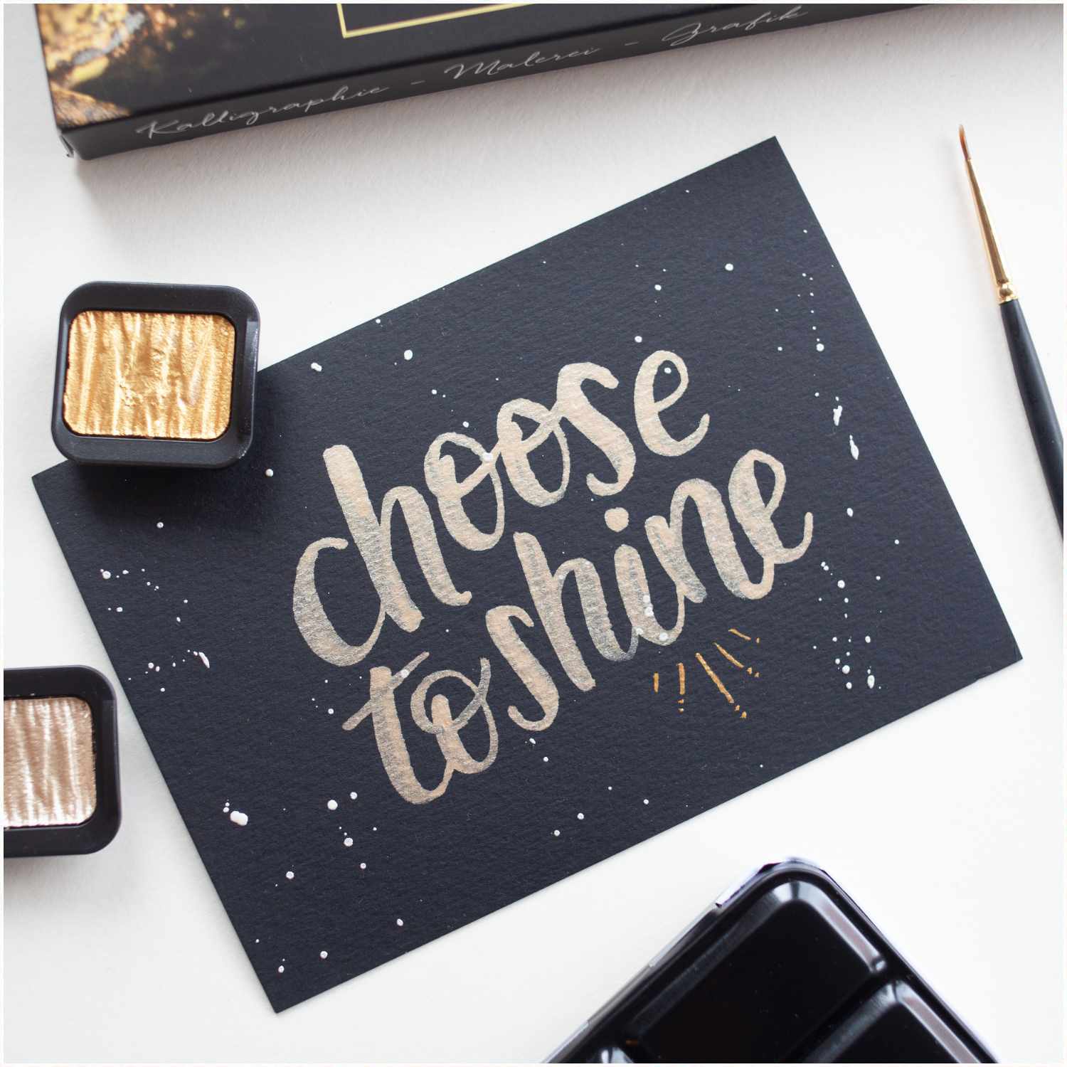 Perlglanzfarben Metals im Metallkasten, präsentiert auf schwarzem Papier mit dem Schriftzug "choose to shine", umgeben von zwei Farbtöpfen und einem Pinsel.