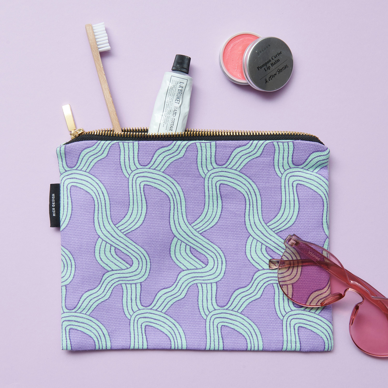 Canvas-Pouches Myzel Flieder: Lila Tasche mit geschwungenem Muster, Zahnbürste, Creme und Sonnenbrille.
