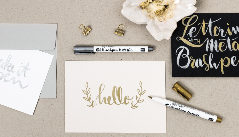 Anleitung Brushlettering: Ein Tisch mit Brushlettering-Stiften, Karten und goldener Dekoration.