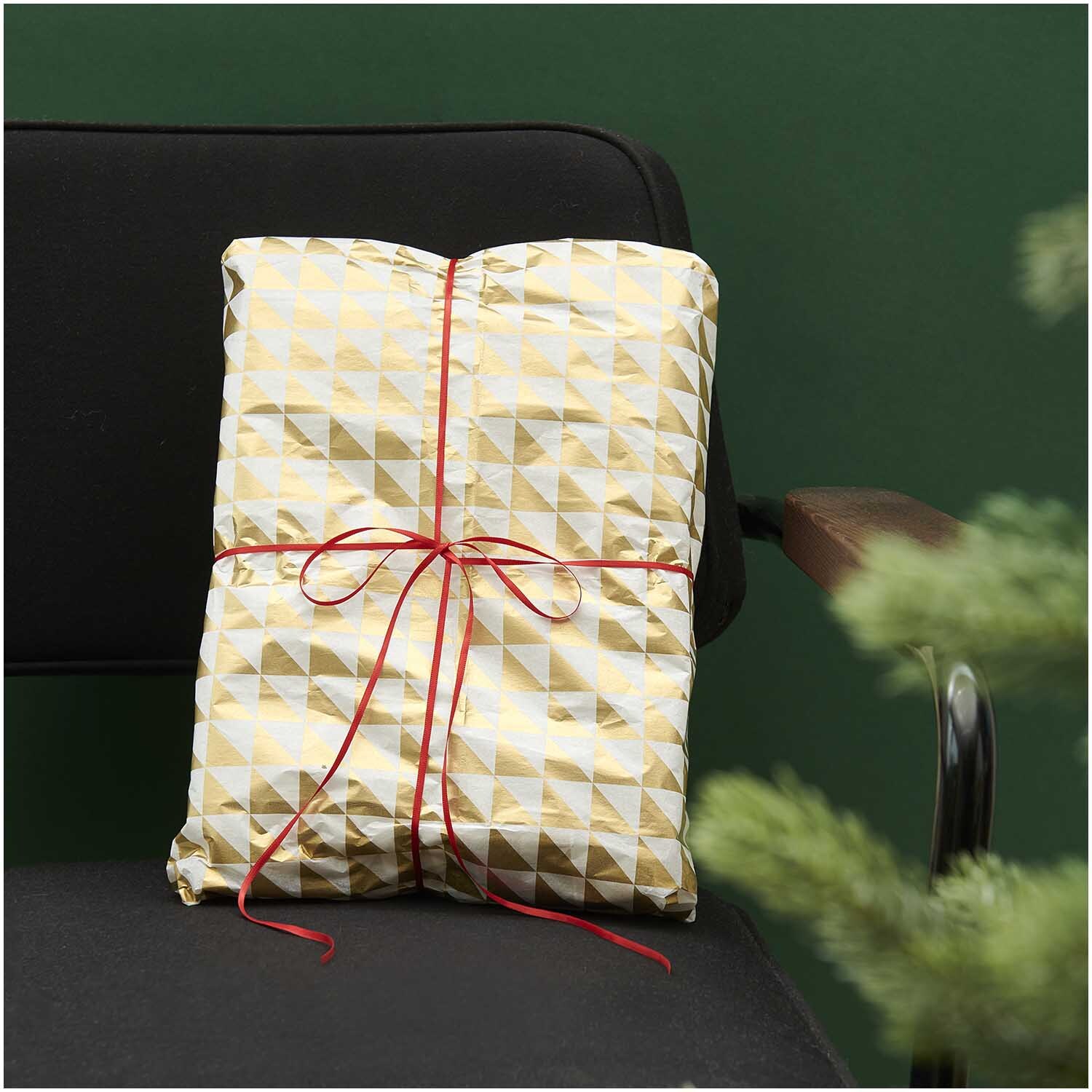 Paper Poetry Seidenpapier mit gold-weißen Dreiecken, dekorativ verpackt mit rotem Band auf einem schwarzen Stuhl. Ideal für stilvolle Geschenke.