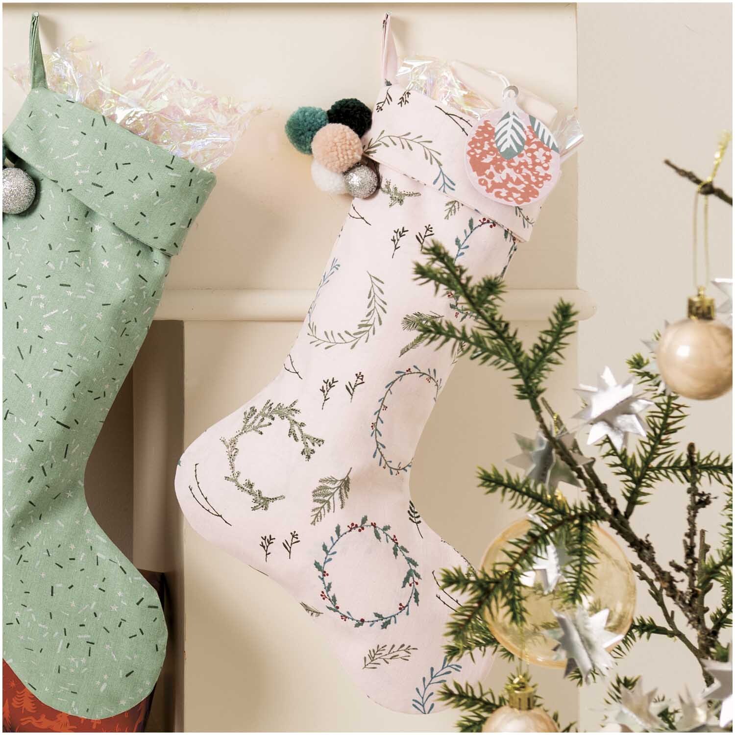 Druckstoff Nostalgic Christmas Kränze in puder-grün und rosa, dekoriert mit weihnachtlichen Motiven, hängt neben einem geschmückten Tannenbaum.
