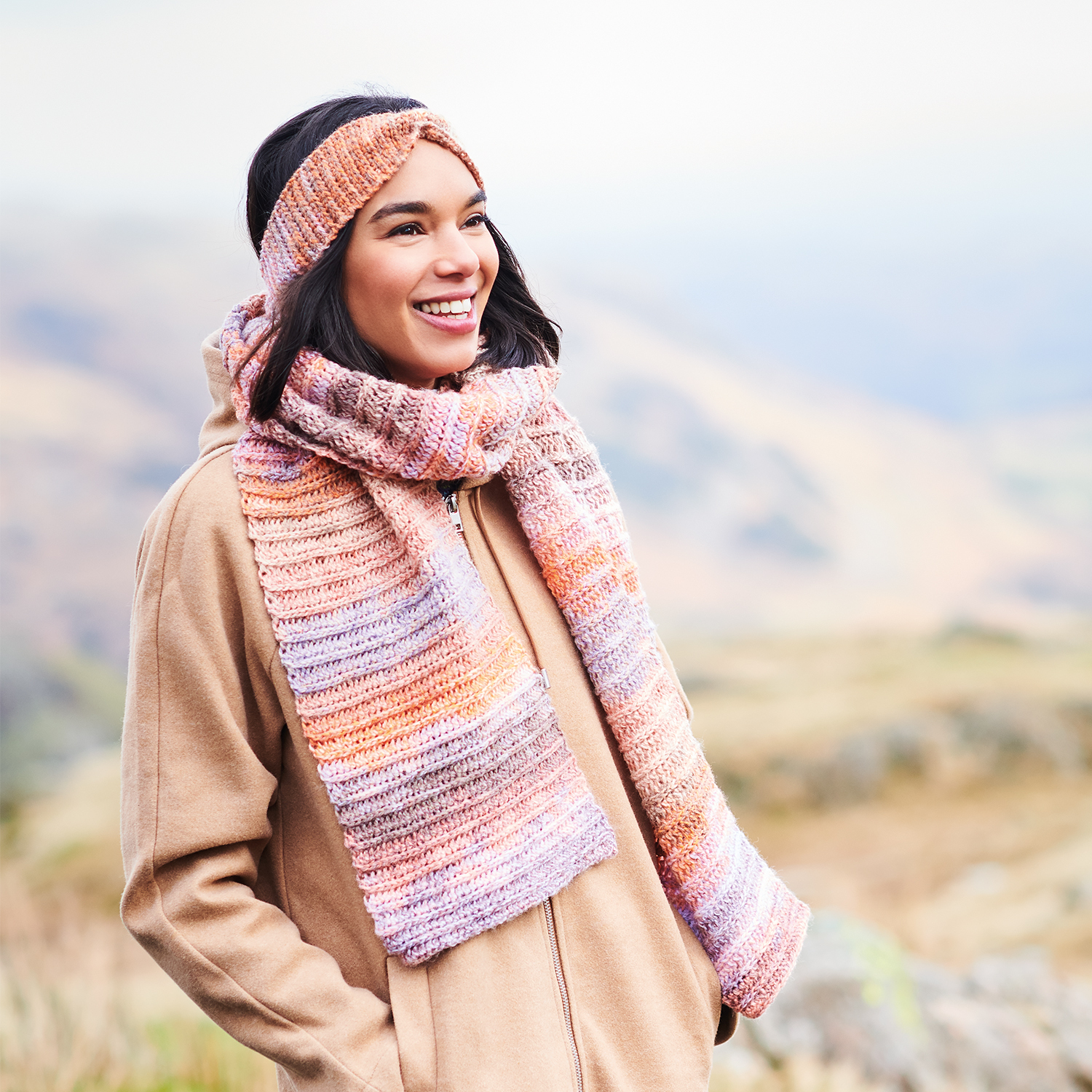 Häkelanleitung Stirnband & Schal aus Creative Chic-Unique dk: Frau trägt gestricktes Set in Pastelltönen, lächelt in natürlicher Landschaft.