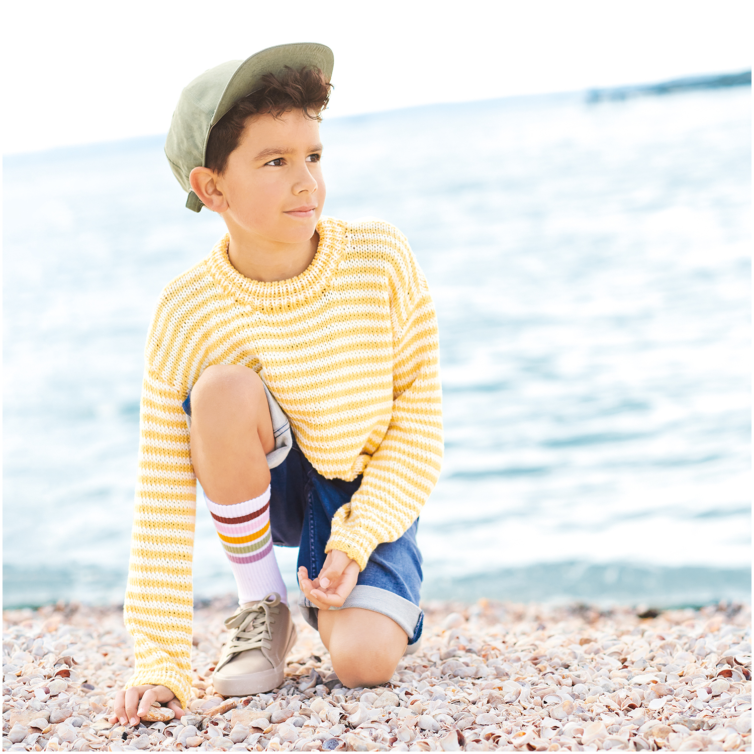 Essentials Organic Cotton aran: Kind in gestreiftem Pullover am Strand, kniend auf Kieselsteinen, mit blauem Meer im Hintergrund.