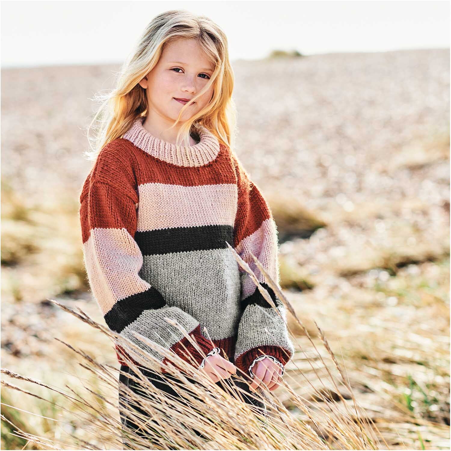 Essentials Super aran: Mädchen in gestreiftem Pullover aus Rico Design Wolle, stehend auf einem Feld.