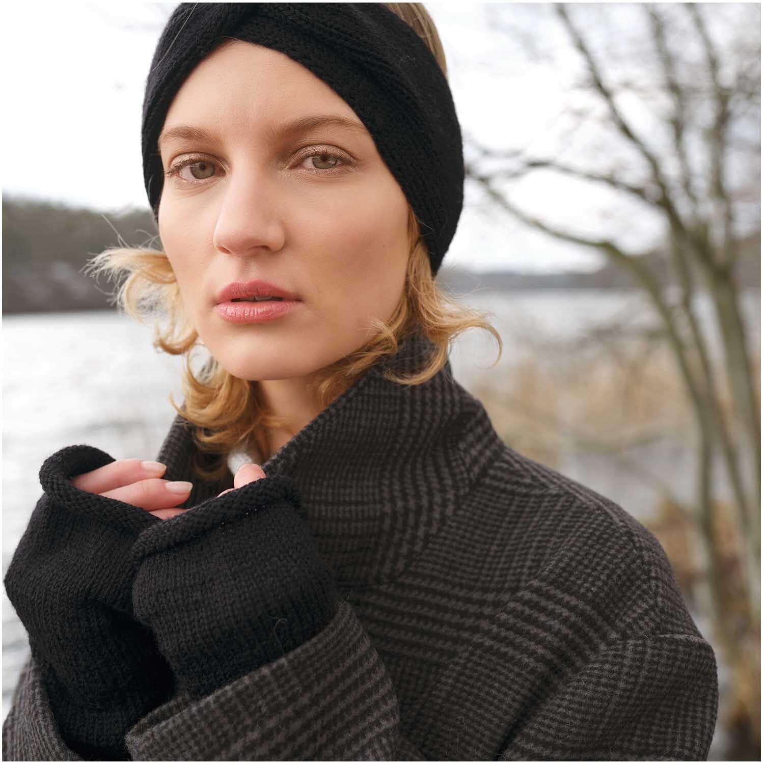 Strickset Stulpen Modell 24: Frau trägt schwarze Stulpen und Stirnband, warm gekleidet am Seeufer.