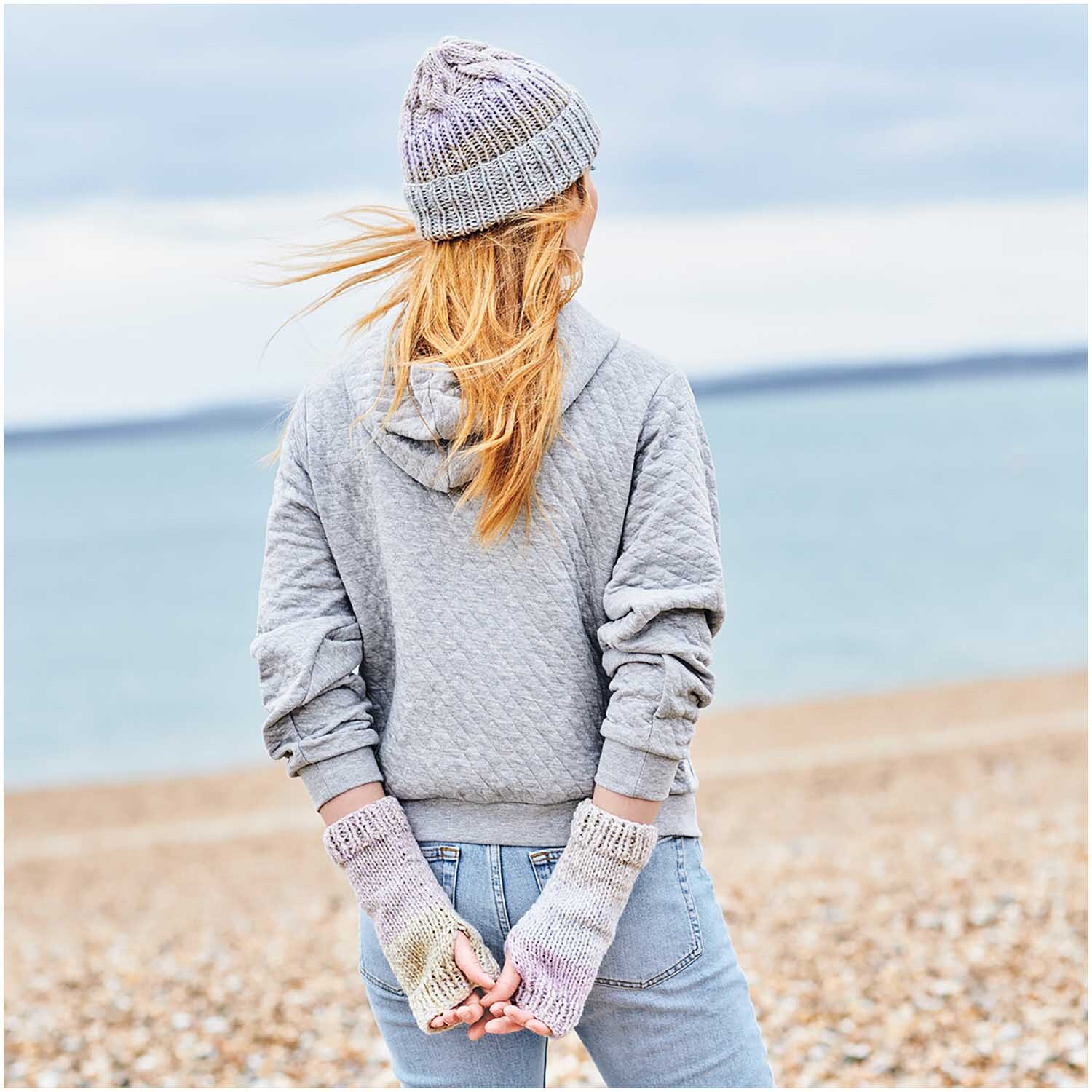 Creative Chic-Unique: Frau am Strand in grauem Hoodie und Mütze, trägt gestrickte Handschuhe von Rico Design.