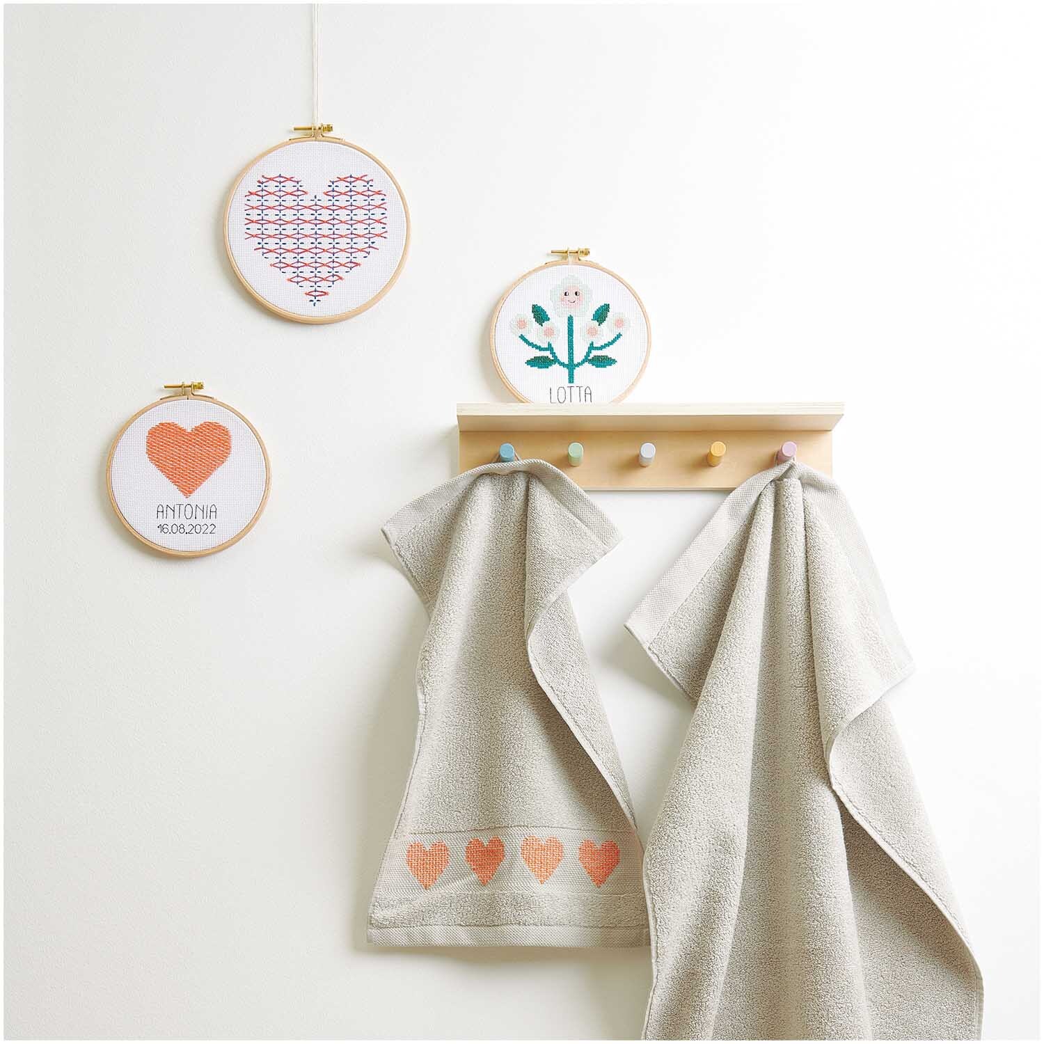 Stickpackung Geburtsbild Herz: Drei Stickrahmen mit Herz- und Blumenmotiven von Rico Design, dekorativ an der Wand über Handtüchern.