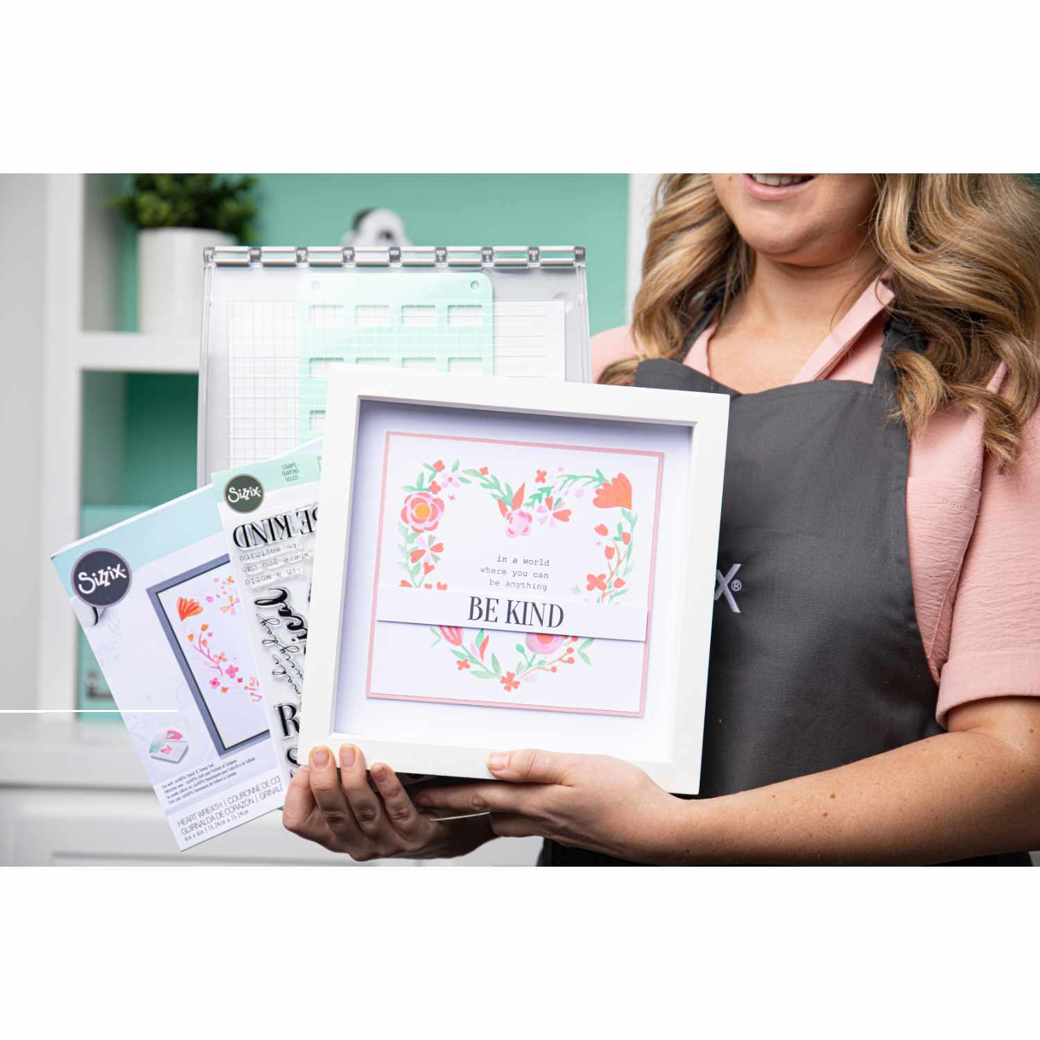 Stencil & Stamp Tool: Frau hält Sizzix-Werkzeug mit floralem Design und "Be Kind"-Botschaft in einem hellen Raum.