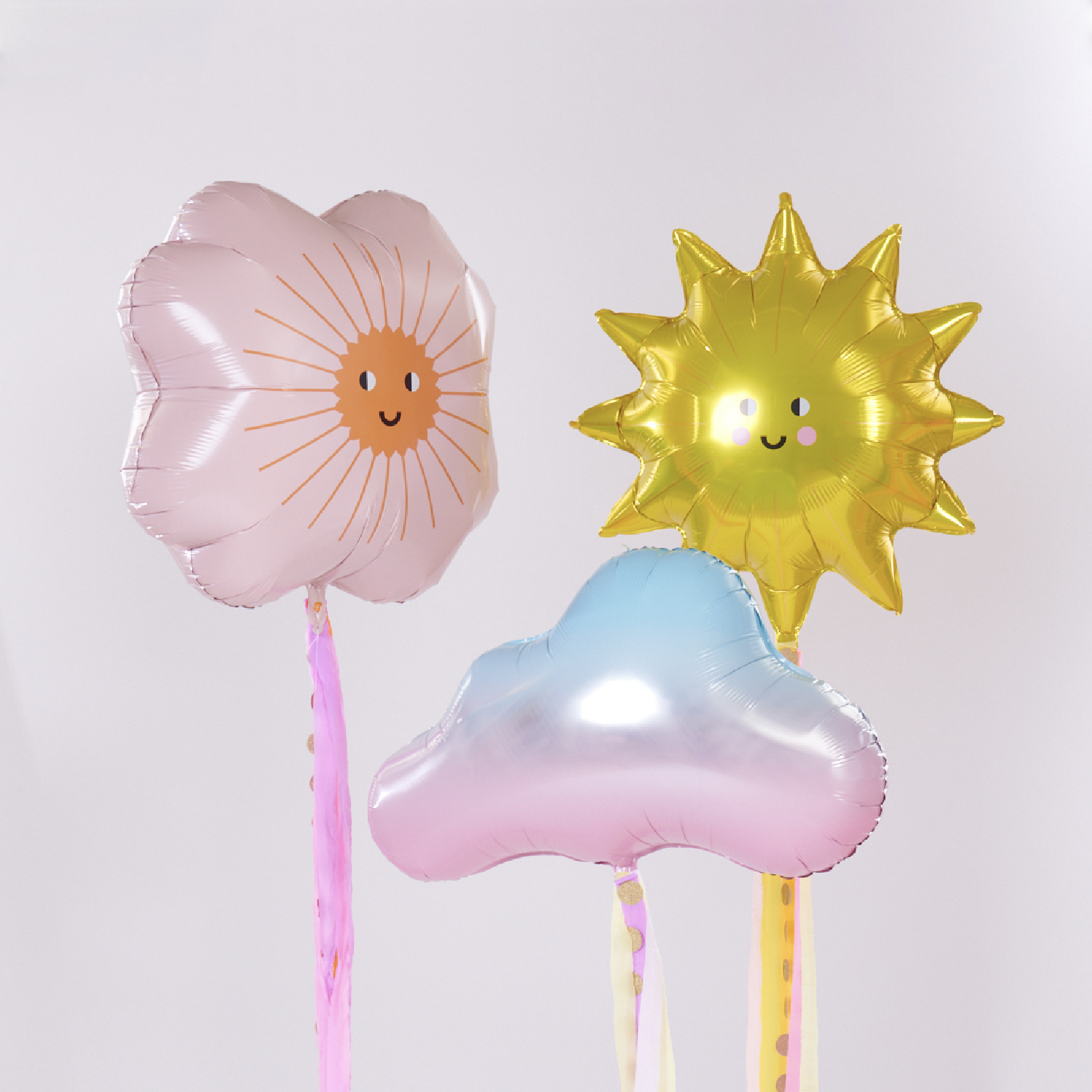 Ein Folienballon in Sonnenform, ein Folienballon in Blumenform und ein Folienballon in Wolkenform, pastellfarben. Fotosetting weiße Wand.