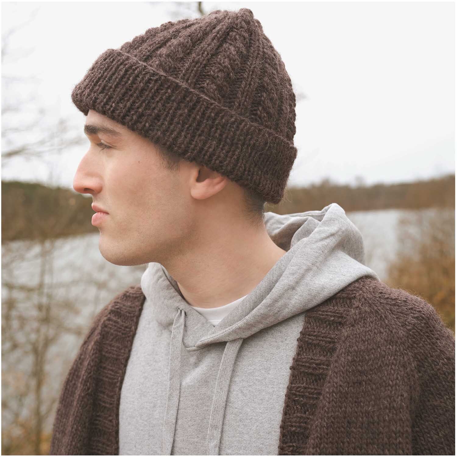 Essentials Organic Wool aran: Mann trägt gestrickte Mütze und Jacke in Braun von Rico Design, im Freien.