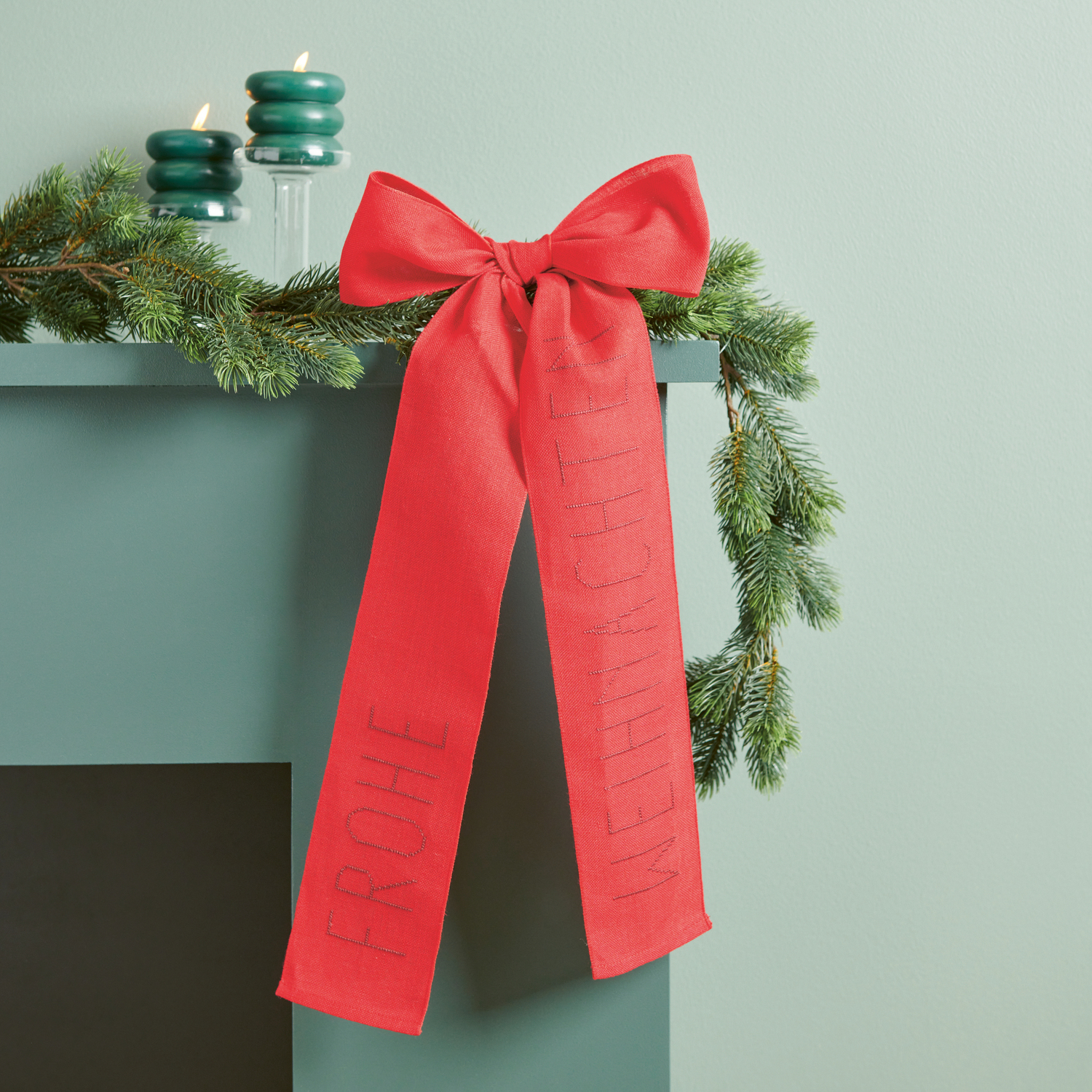 Stickbuch Put a Bow on It Nr. 185: Rote Schleife mit "Frohe Weihnachten" auf grünem Kamin mit Tannenzweigen.
