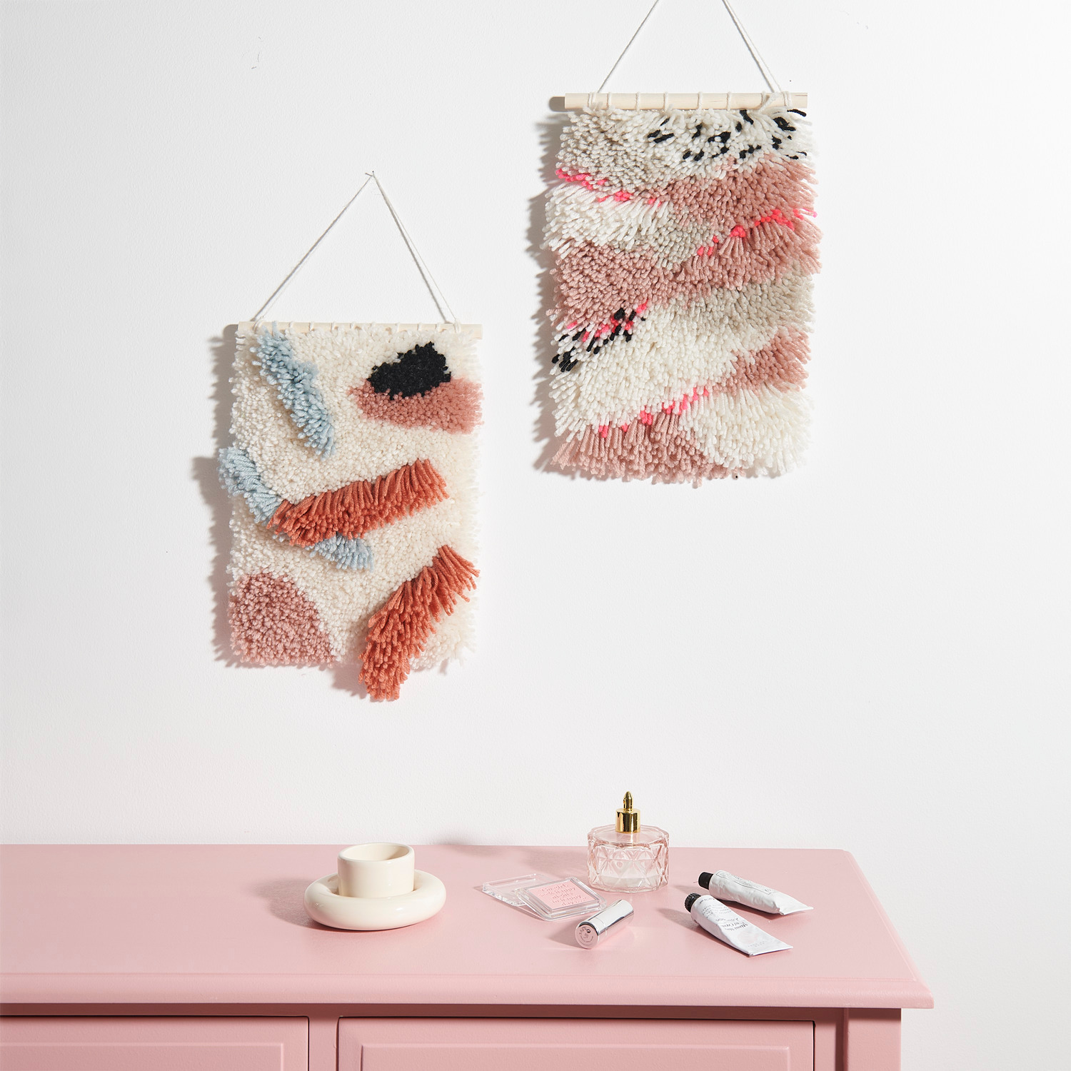 Rico Hooki Hooki Knüpfset Wandbehang Neon Landschaft zeigt zwei bunte, texturierte Wandbehänge über einer rosa Kommode.