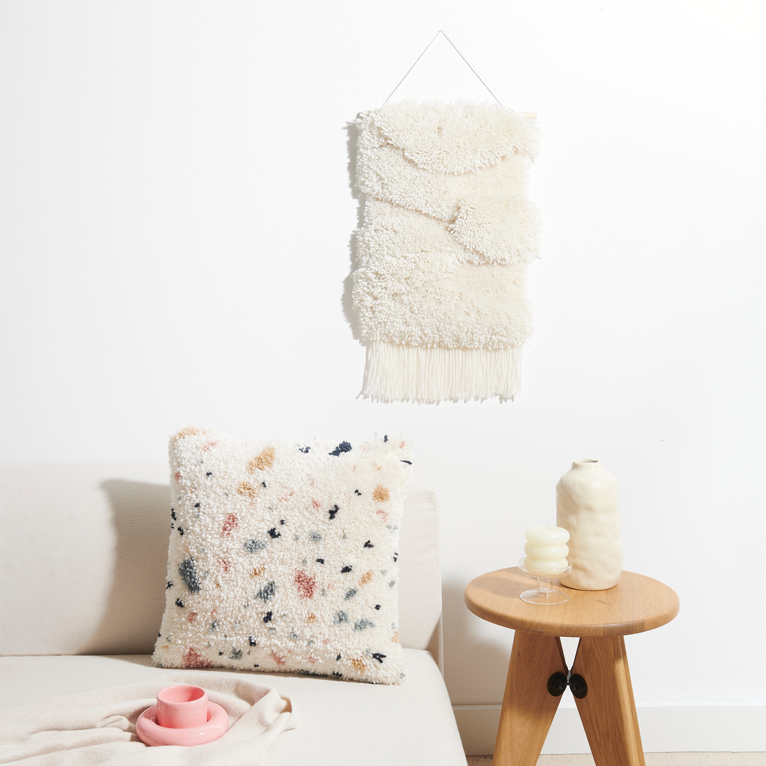 Rico Hooki Hooki Knüpfset Wandbehang Weiße Landschaft: Weißer Wandbehang über Sofa mit gemustertem Kissen, dekoriert mit Tisch und Vase.