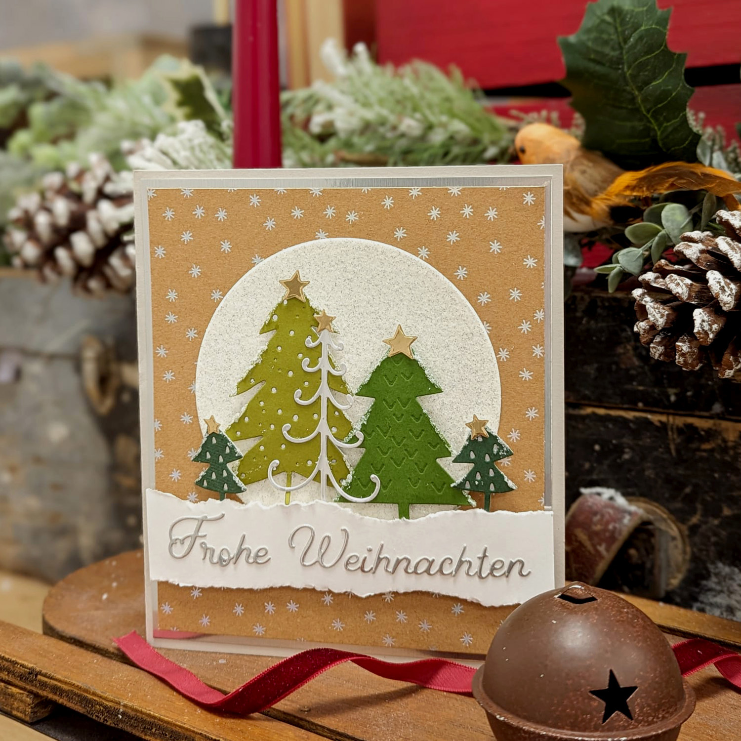 Bastelanleitung Weihnachtskarte mit der Sizzix gestalten: Handgemachte Karte mit Tannenbäumen und "Frohe Weihnachten" Schriftzug, umgeben von weihnachtlicher Dekoration.