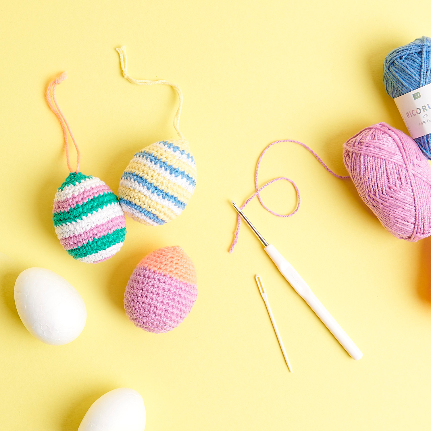 Häkelset Ricorumi Ostereier classic: Bunte, gehäkelte Ostereier mit Garn und Häkelnadel auf gelbem Hintergrund.