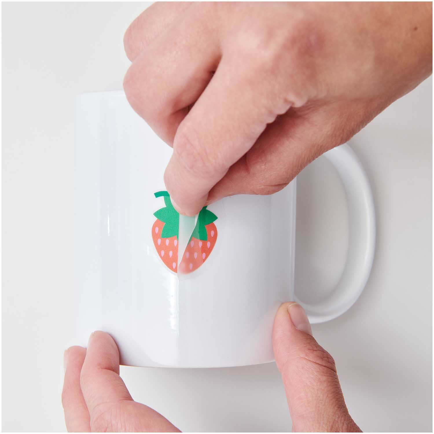 Transfermotiv Früchte & Blumen: Hände kleben Erdbeer-Aufkleber auf weiße Tasse. Set enthält 15 Stück von Rico Design.