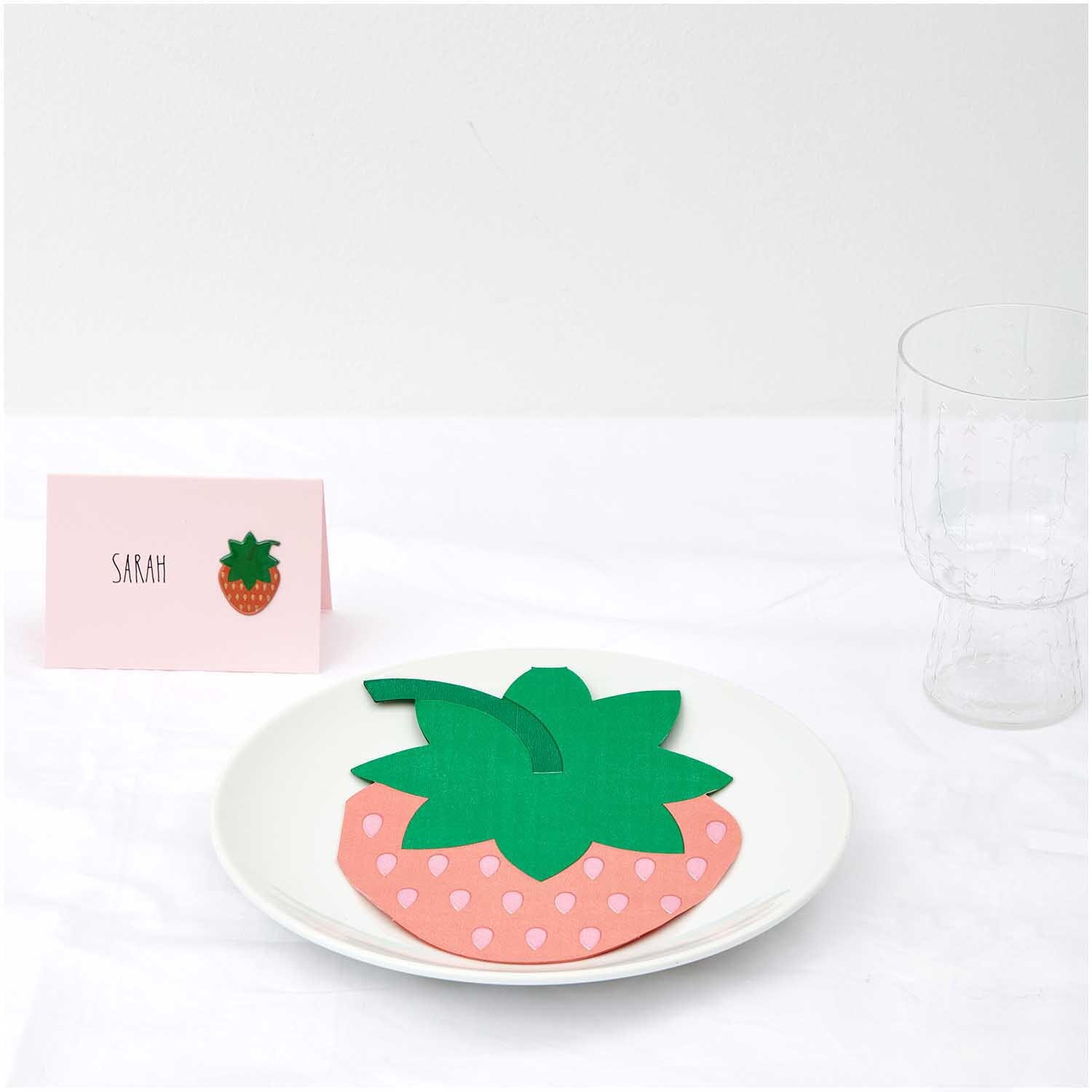 Servietten Erdbeere 12x16cm 20 Stück: Erdbeerförmige Servietten auf einem Teller, dekorativ arrangiert neben einem Namenskärtchen. Ideal für sommerliche Anlässe.