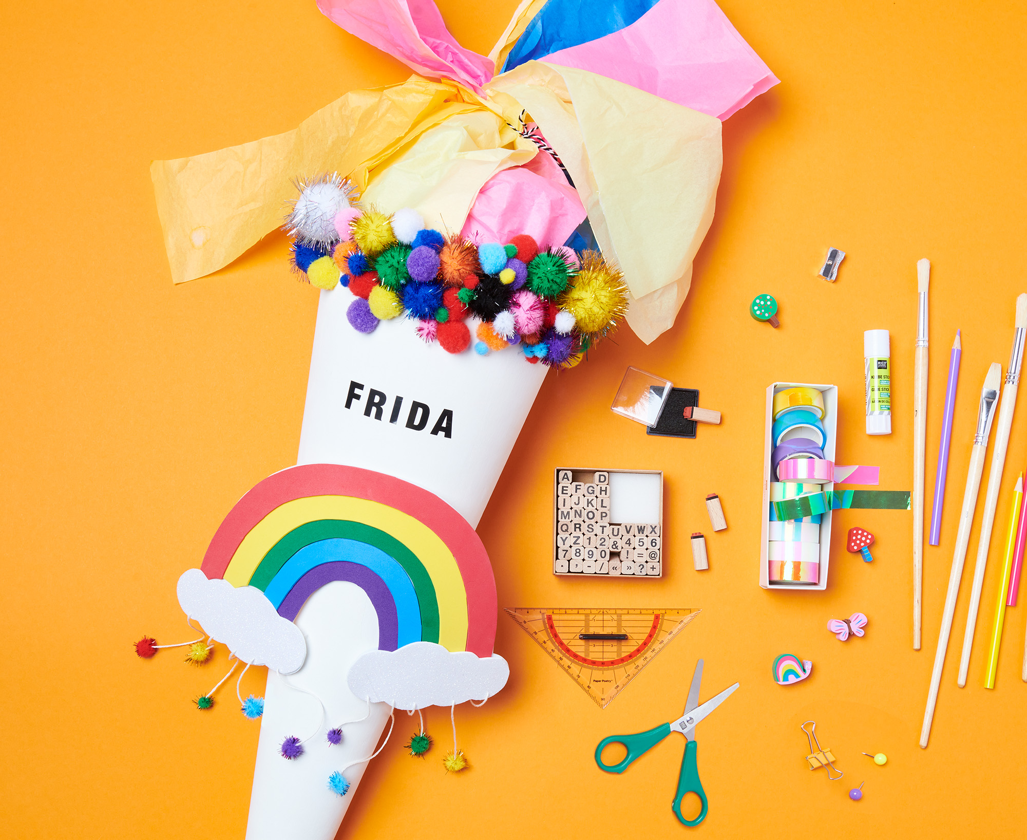 Weiße Schultüte mit Regenbogen und bunten Pompons, daneben liegen Stifte, Pinsel, Stempel, bunte Tapes, Radiergummis, ein Lineal und eine Schere. Flatlay von Produkten.