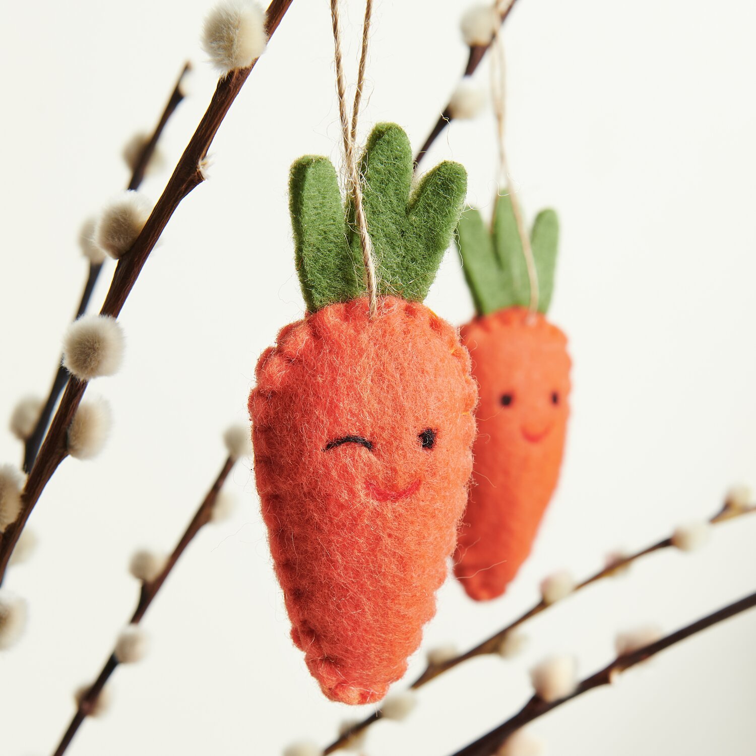 Aufhänger Filz-Möhre Zwinkernd 2D: Zwei orange Filz-Möhren mit Gesicht, hängend an Zweigen.