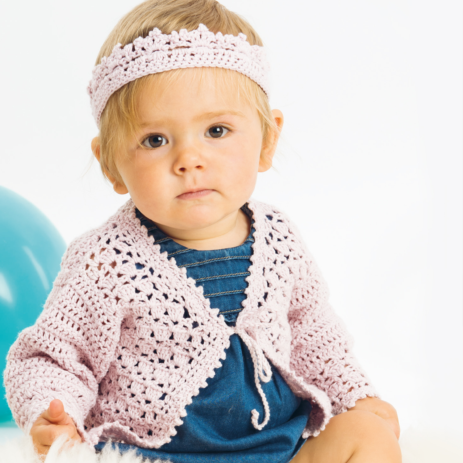 Häkelanleitung Baby-Jacke mit Krone: Baby trägt rosa gehäkelte Jacke und Stirnband, sitzt vor blauem Ball.