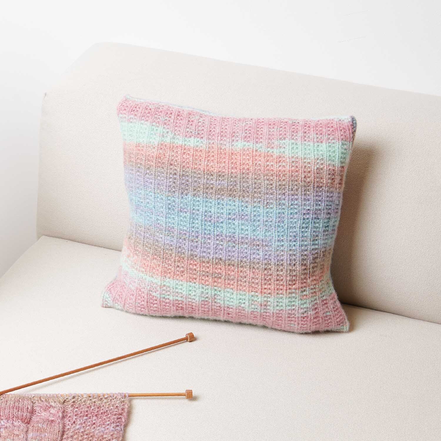 Strickanleitung Kissen aus Creative Painted Power: Pastellfarbenes Kissen auf beigem Sofa, gestrickt mit buntem Garn.