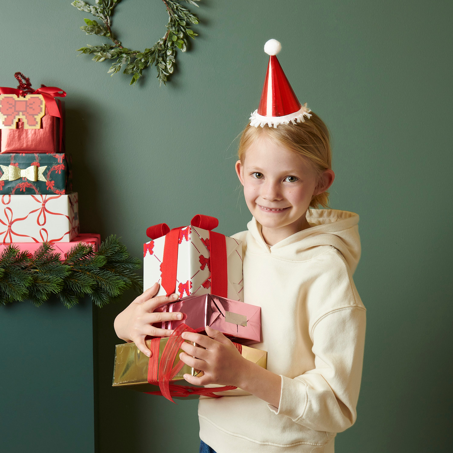 Mini-Partyhüte Weihnachtsmann: Kind mit roten Hüten von Rico Design, hält Geschenke vor grünem Hintergrund. Ø8x14cm 8 Stück.