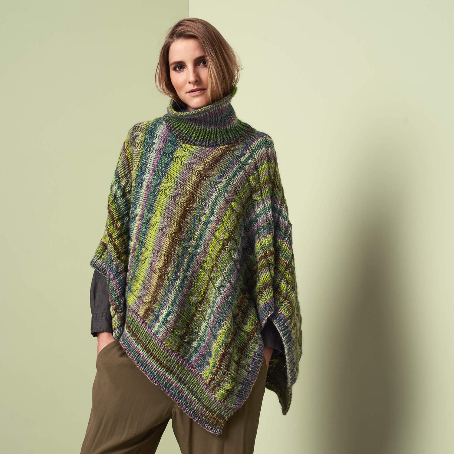 Strickanleitung Poncho aus Rigato: Frau trägt bunten, gestreiften Poncho mit Rollkragen vor neutralem Hintergrund.