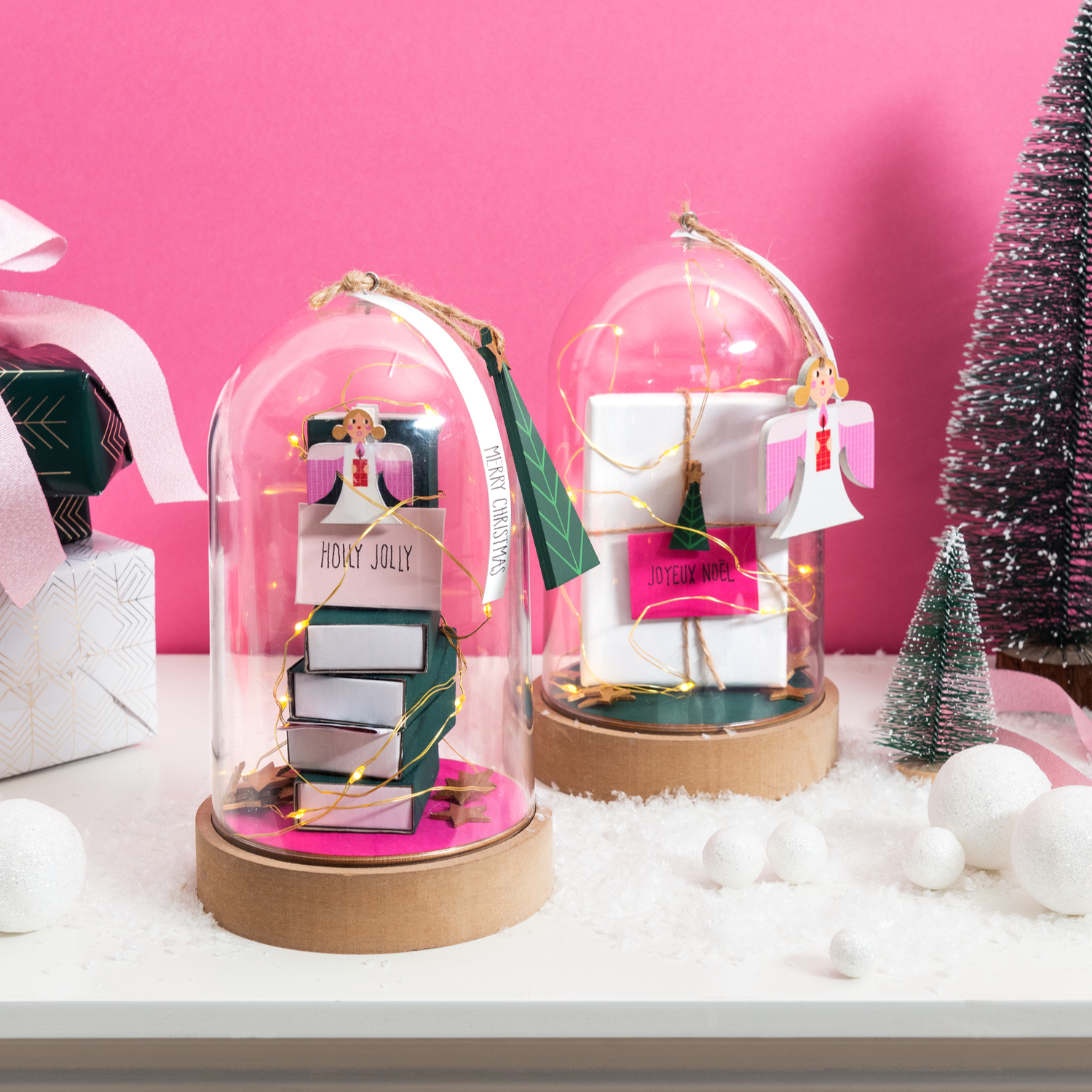 Bastelanleitung in Dekohaube: Zwei dekorative Glashauben mit Geschenken und festlichen Anhängern auf einem Tisch vor rosa Hintergrund.