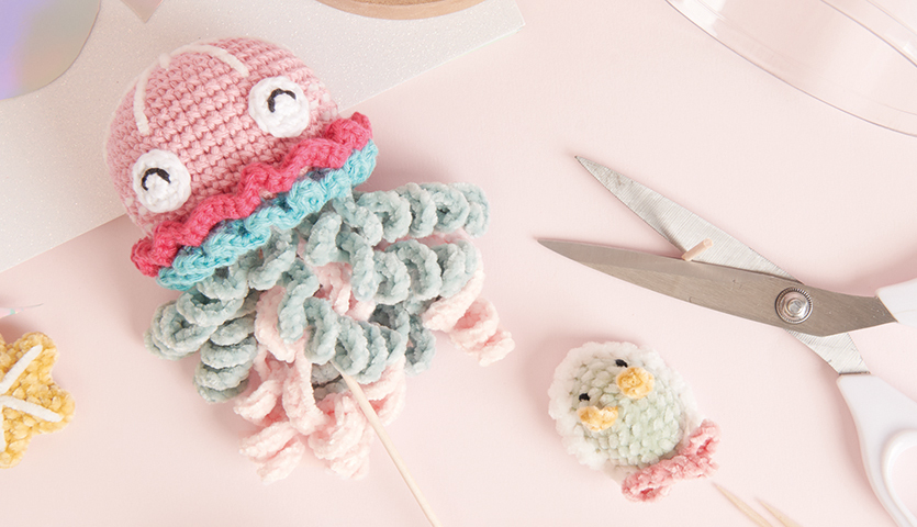 Häkelanleitung Qualle aus Ricorumi und Ricorumi Nilli Nilli, pastellfarbene gehäkelte Qualle mit rosa, blau und weiß, neben Schere und kleinem Vogel.