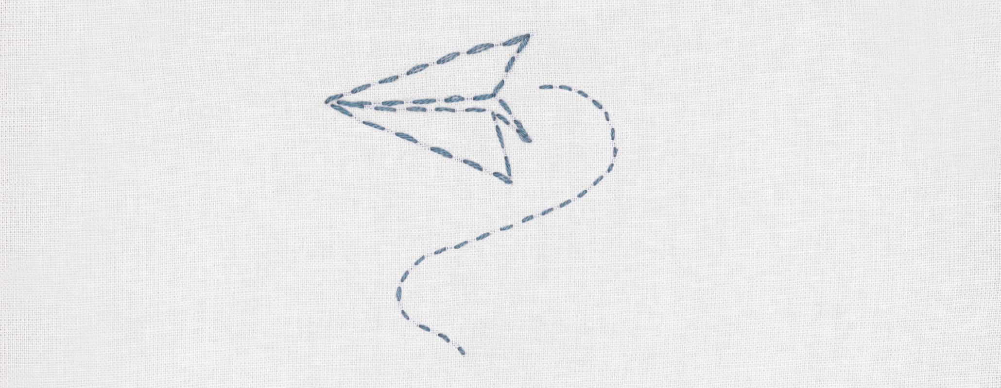 Der Umriss eines blauen Papierfliegers ist im Vorstich auf weißen Stoff gestickt
