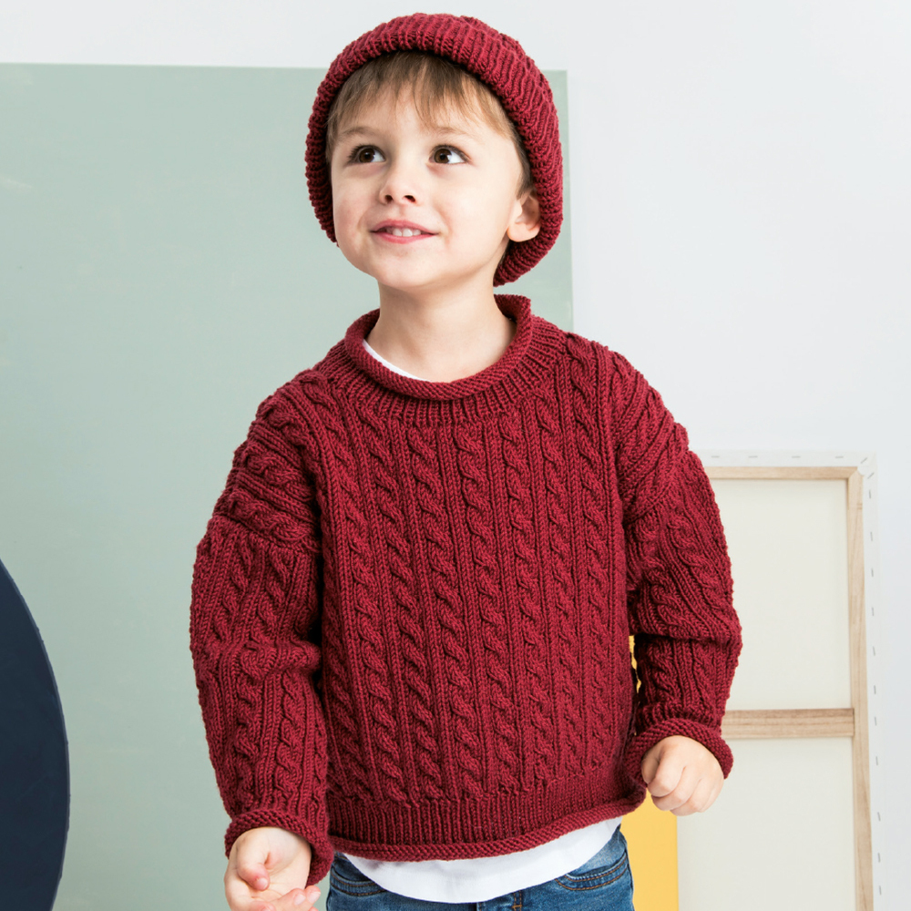 Strickanleitung Zopfpullover mit passender Mütze: Kind trägt roten Zopfpullover und Mütze, lächelnd.