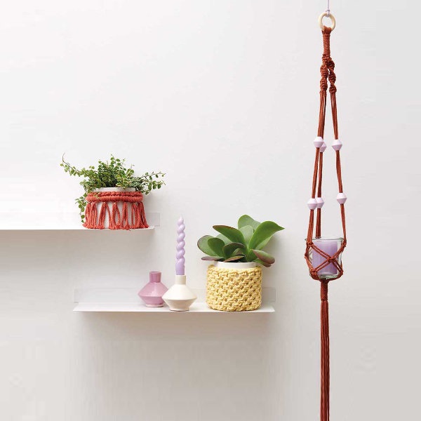 Zwei Wandregale mit unterschiedlich umhäkelten Blumentöpfen und zwei Kerzenhaltern, daneben hängt eine rötliche Blumenampel mit Perlen und einem Glas.