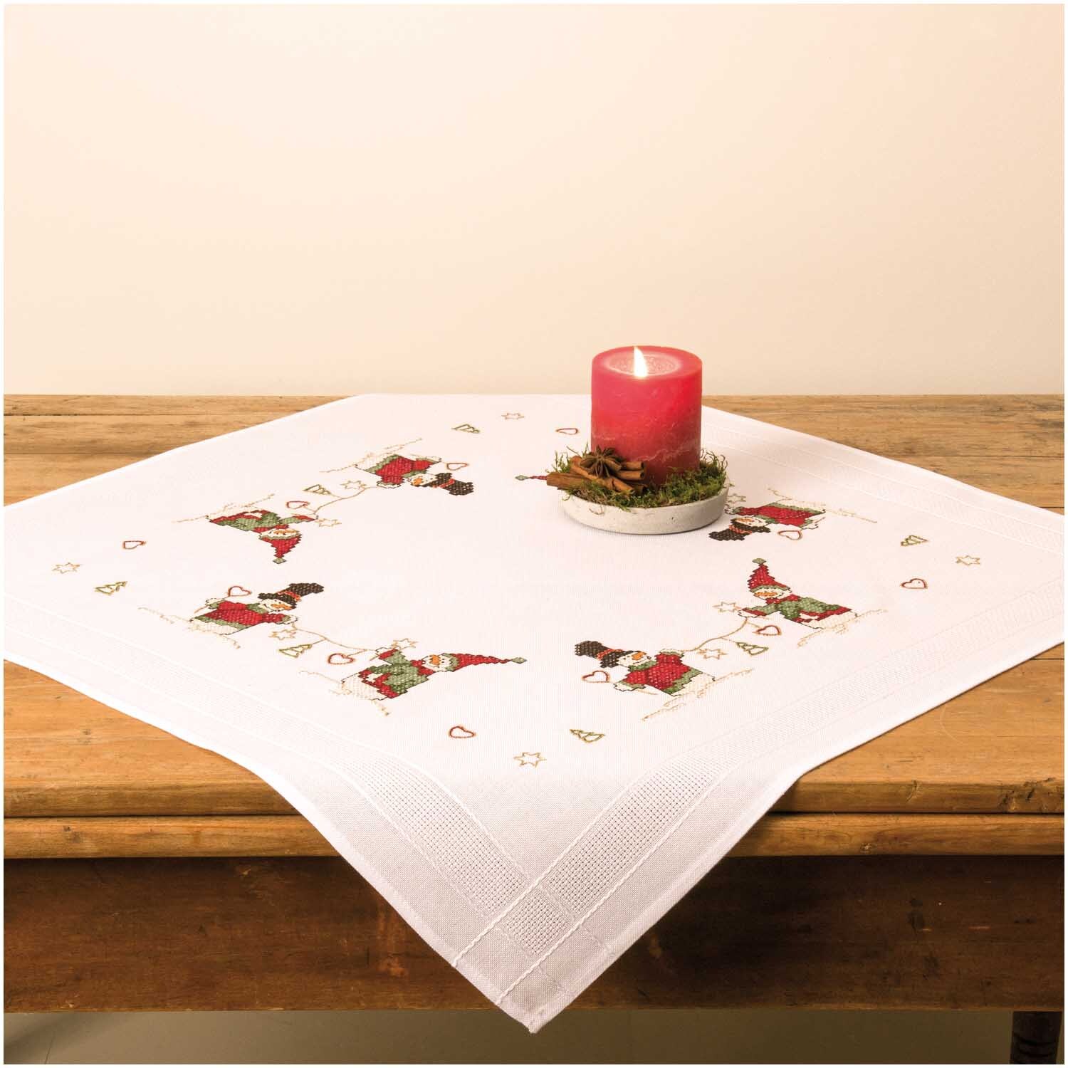 Stickpackung Tischdecke Schneemänner 80x80cm zeigt eine weiße Tischdecke mit Schneemann-Motiven und einer roten Kerze in der Mitte.