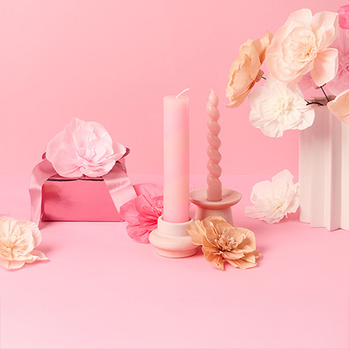 Zwei rosa Kerzenhalter mit passenden rosa Kerzen, drumherum Papierblüten und ein rosa verpacktes Geschenk. Produkte aus der Kollektion La Vie En Rose.