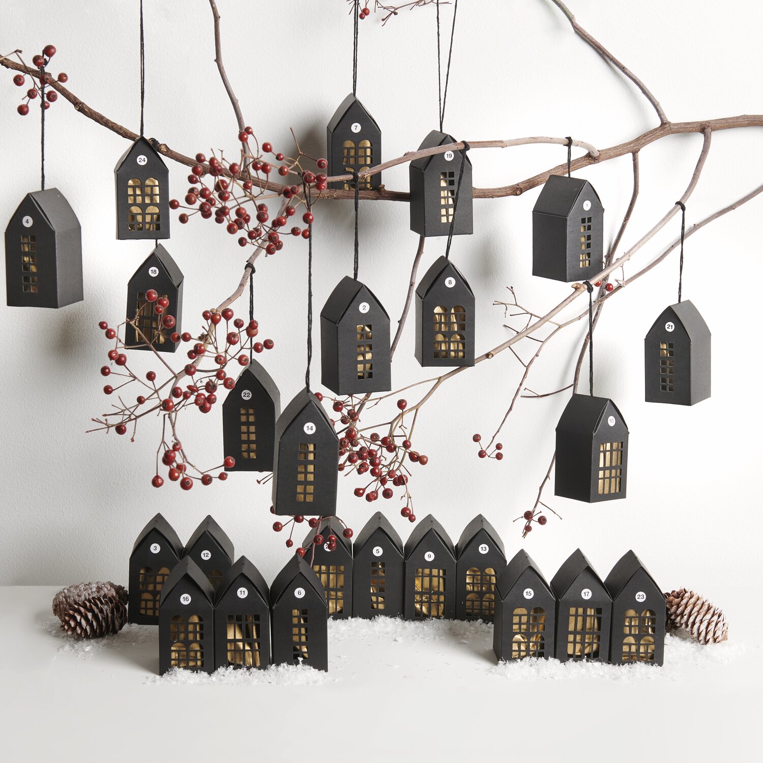 Adventskalender Papphäuser 24 Stück: Schwarze Häuschen von Rico Design, dekorativ an Zweigen hängend, mit roten Beeren und Tannenzapfen.