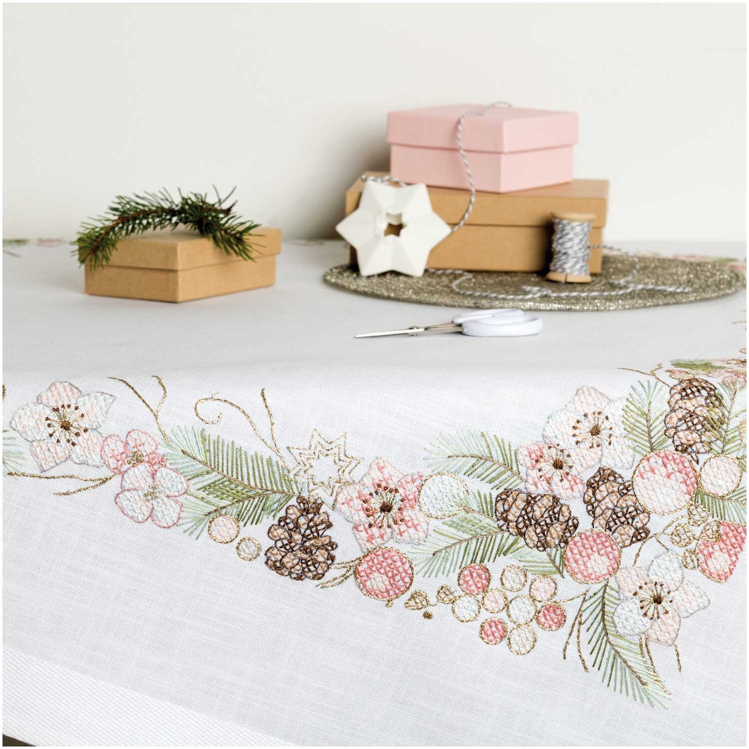Stickpackung Tischdecke Christrosen 90x90cm von Rico Design zeigt bestickte Blumen und Tannenzapfen auf weißem Stoff, umgeben von Geschenkboxen.