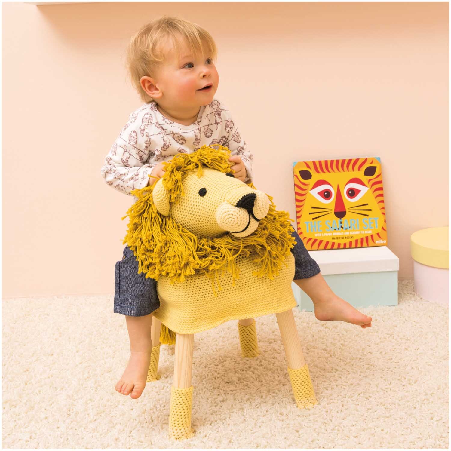 Creative Cotton aran: Ein Kind sitzt auf einem Hocker mit gestricktem Löwenbezug in Gelb, umgeben von buntem Spielzeug.