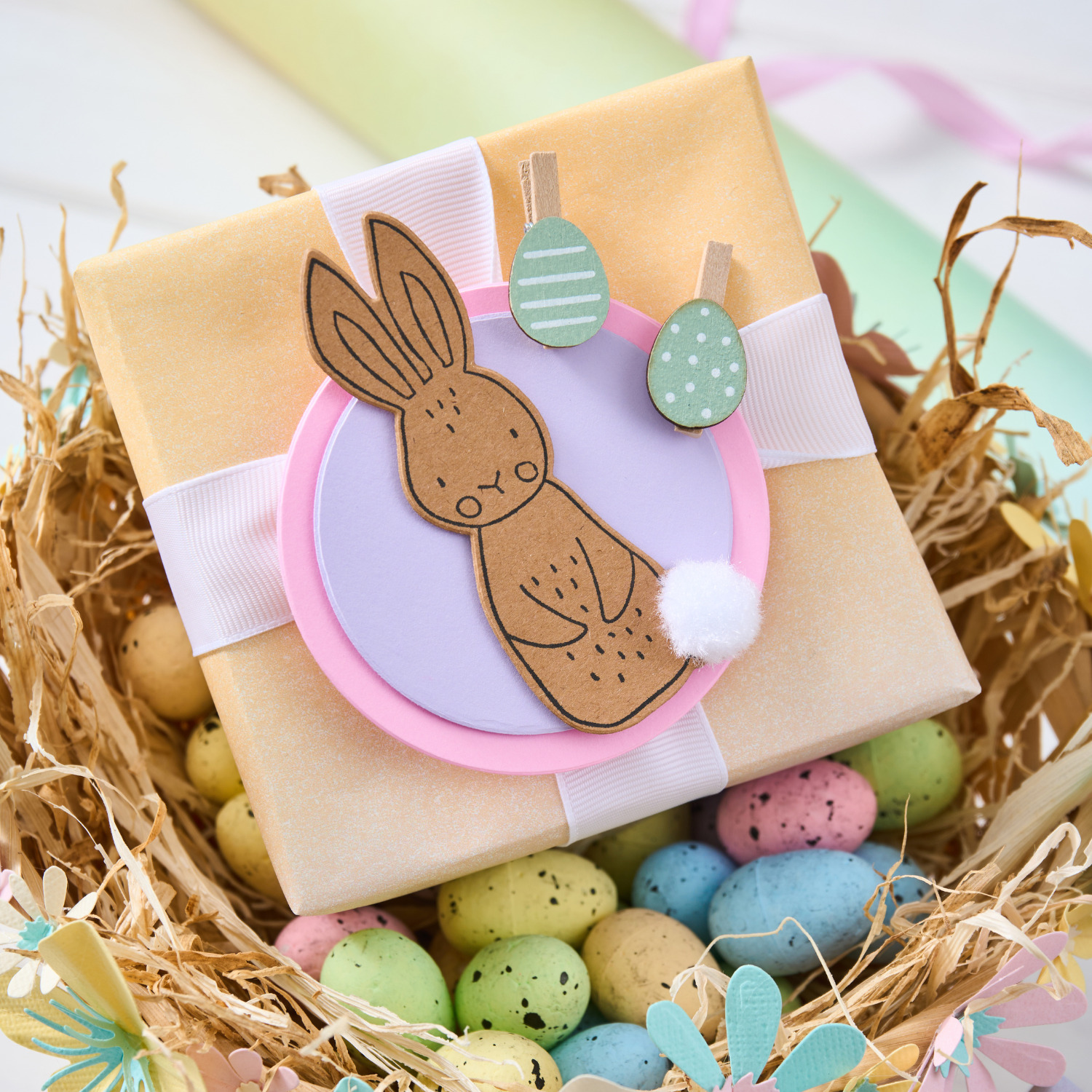 Bastelanleitung Osterhasen-Geschenkanhänger mit Sizzix: Pastellfarbenes Geschenk im Nest, dekoriert mit Hasenmotiv und Ostereiern.