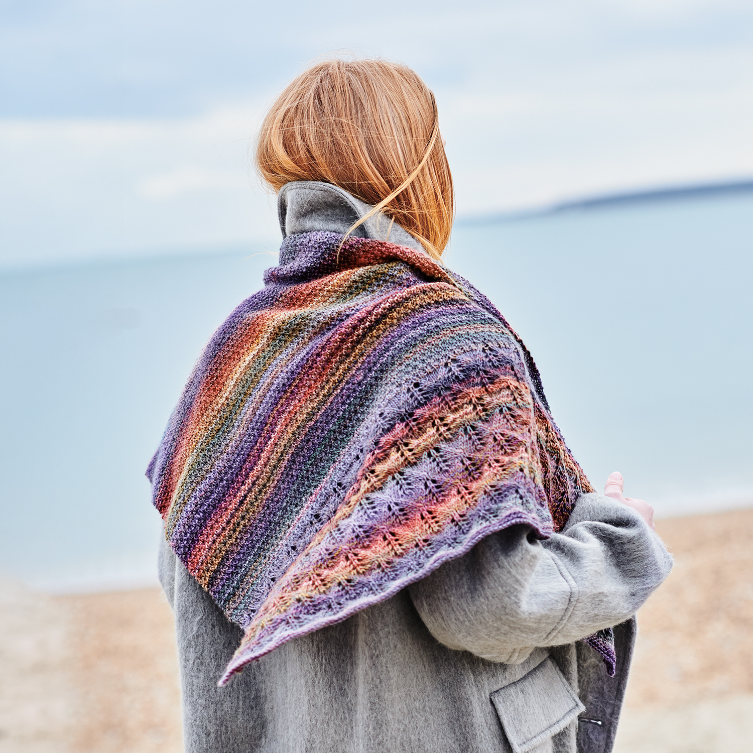 Strickanleitung Tuch aus Creative Melange aran Wonderball: Frau am Strand mit buntem, gestricktem Tuch über grauem Mantel.